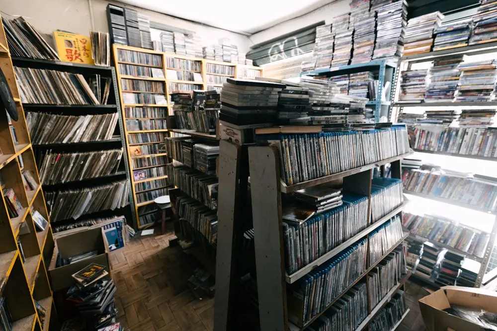 Na imagem, uma imagem de ângulo aberto mostrando o interior abarrotado de um sebo ou loja de mídia usada. O espaço é preenchido com estantes de madeira e metal, todas superlotadas. No lado esquerdo, estantes de madeira contêm pilhas de discos de vinil ou CDs. No centro, uma estante de chão, possivelmente de livros e DVDs ou Blu-rays, domina o primeiro plano, com itens empilhados no topo. À direita e ao fundo, estantes de metal azul claro e escuro estão repletas de livros e pilhas de mídia em caixas finas. O chão é de tábuas de madeira ou parquê. A luz natural entra pela parte superior direita, possivelmente vinda de uma janela ou porta entreaberta, realçando a densidade e a desordem organizada do inventário.