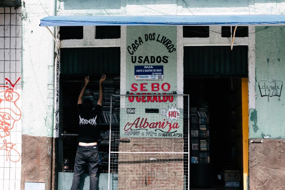 Na imagem, a fachada de um sebo (livraria de livros usados). O prédio é antigo, com a parte superior pintada em um verde ou azul claro desbotado e a parte inferior em pedra marrom. Um toldo azul se estende por cima da entrada. No centro da fachada, um grande cartaz fixo exibe: 