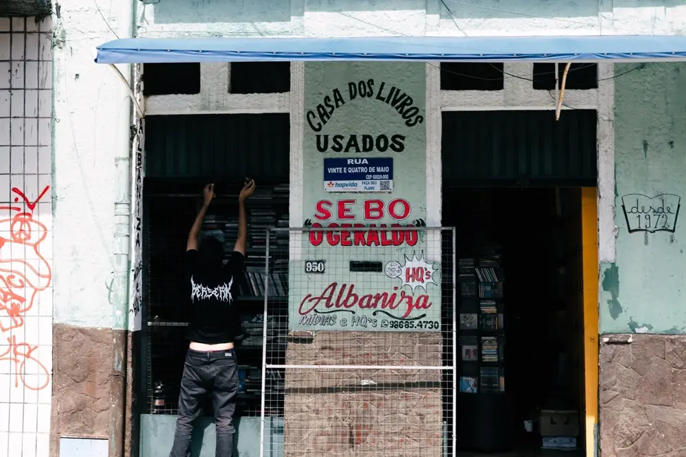 Na imagem, a fachada de um sebo (livraria de livros usados). O prédio é antigo, com a parte superior pintada em um verde ou azul claro desbotado e a parte inferior em pedra marrom. Um toldo azul se estende por cima da entrada. No centro da fachada, um grande cartaz fixo exibe: 