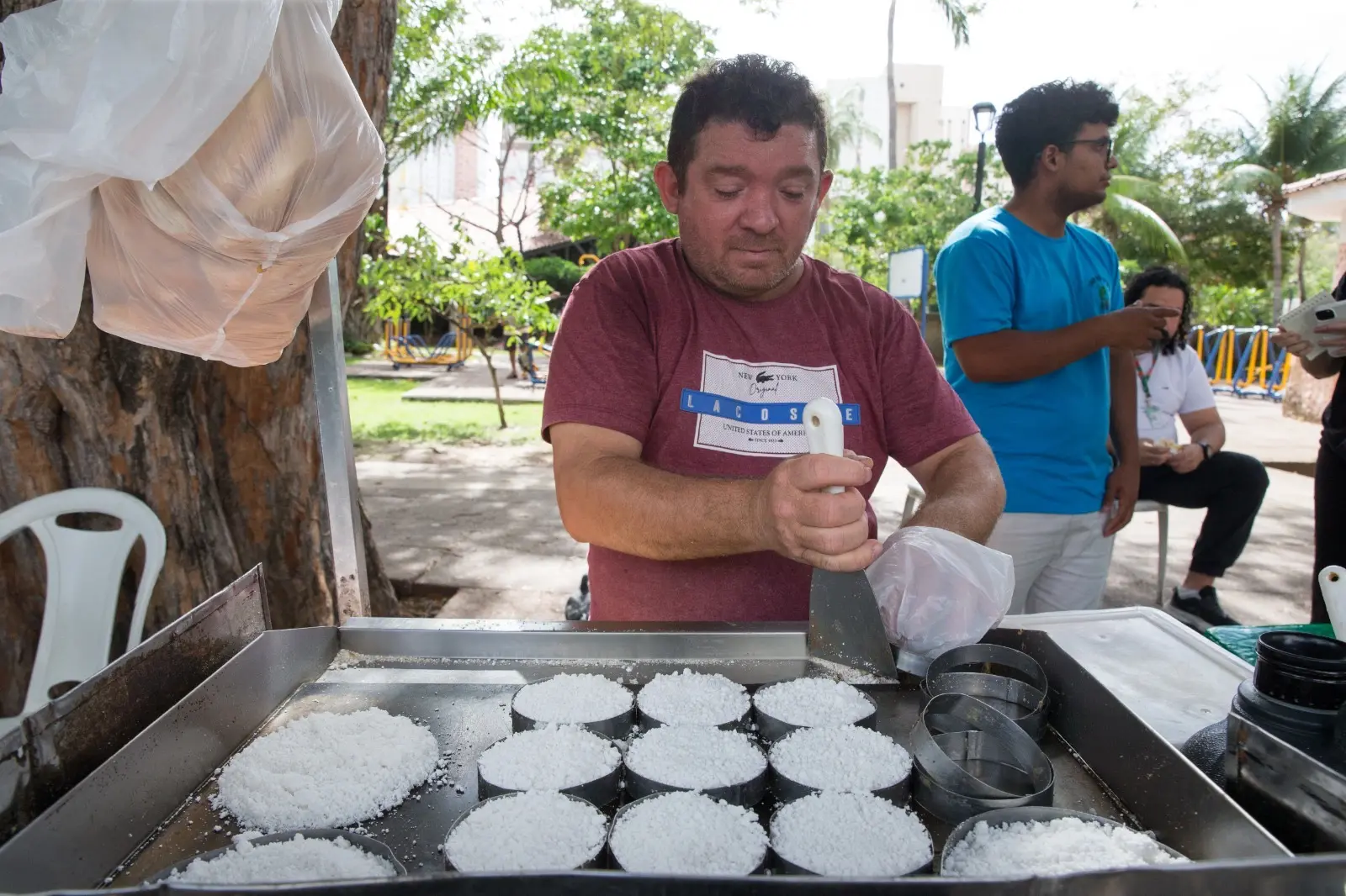Homem branco de camisa vermelha com detalhes branco e azul preparando tapiocas redondas tradicionais em uma praça, com árvores verdes ao fundo e um homem negro de blusa azul olhando para o lado.