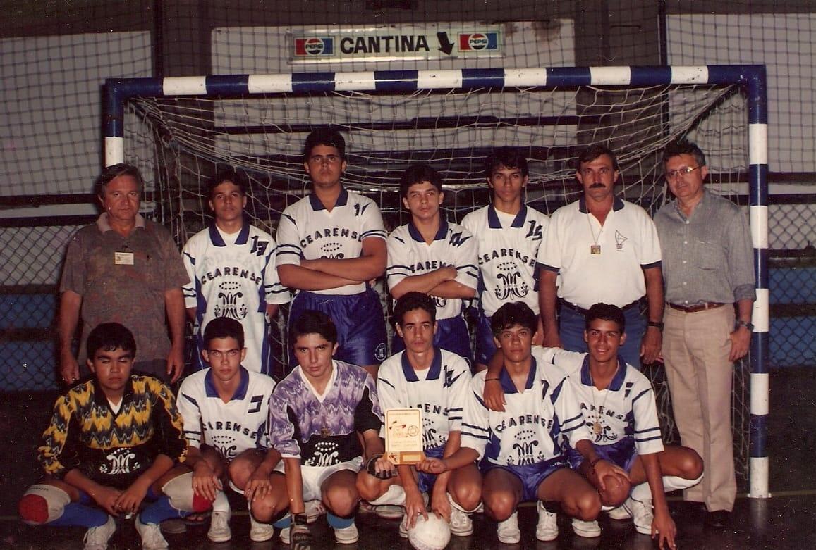 Foto de time de futsal dos anos 90, 12 pessoas (jovens e adultos) em uniformes brancos e azuis posando com troféu e bola. O fundo mostra um gol e um letreiro de CANTINA com logo Pepsi.