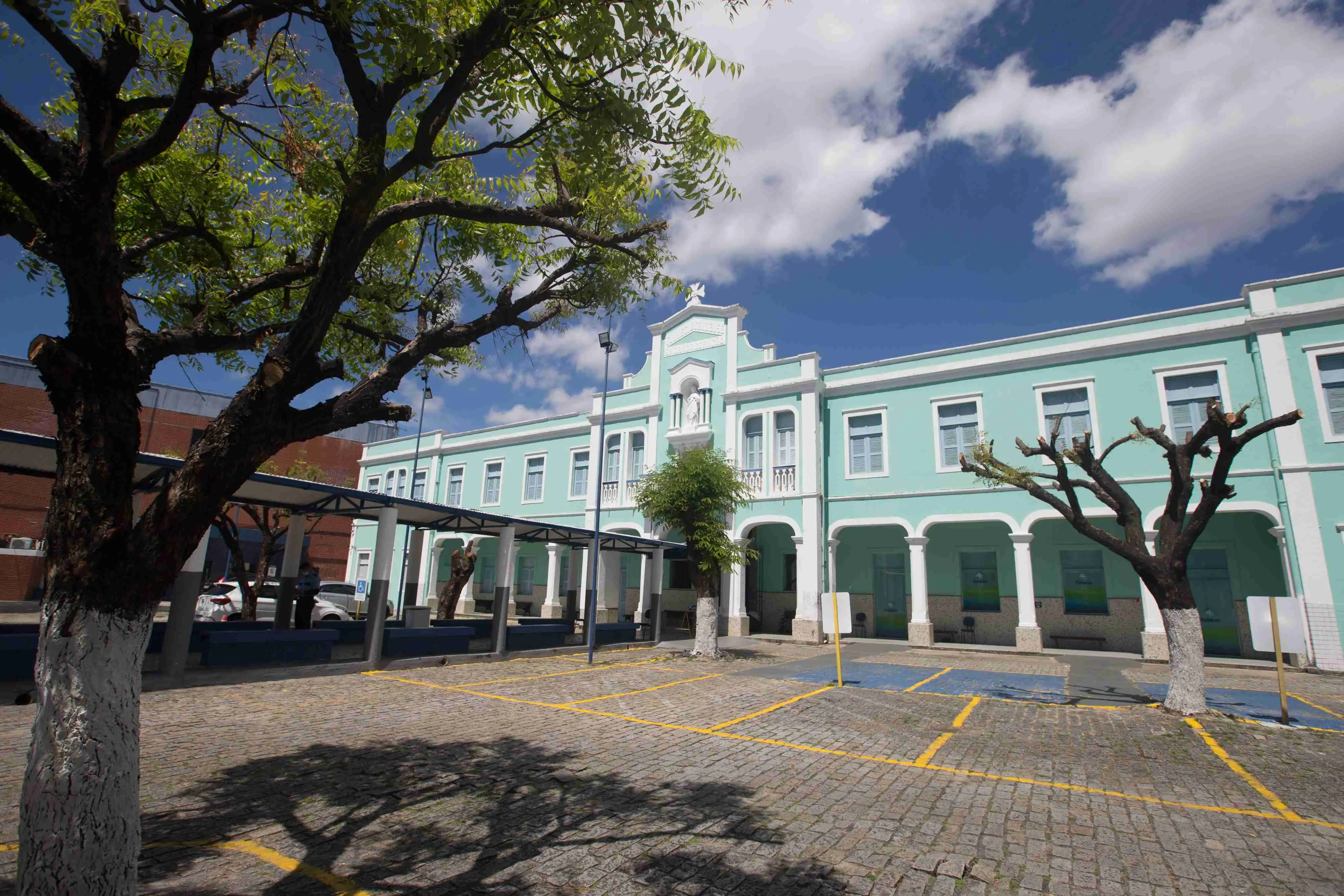 Fachada principal do Colégio Marista de Fortaleza. O prédio, em estilo colonial/eclético, é pintado de verde-água e branco, com arcos e uma imagem religiosa no topo. O pátio de paralelepípedos tem vagas de estacionamento. Duas árvores em primeiro plano emolduram o edifício sob um céu azul.