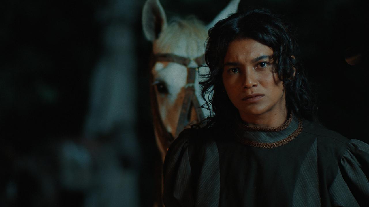 Foto escura e dramática da atriz Dira Paes, de cabelos longos e escuros, no filme 'Lua Cambará'. Ela está vestindo uma roupa de época escura com um detalhe de gola dourada, olhando para a frente com uma expressão séria. Ao fundo, fora de foco e ligeiramente desfocado, está a cabeça de um cavalo branco ou de cor clara. A iluminação é baixa, criando uma atmosfera noturna ou sombria.