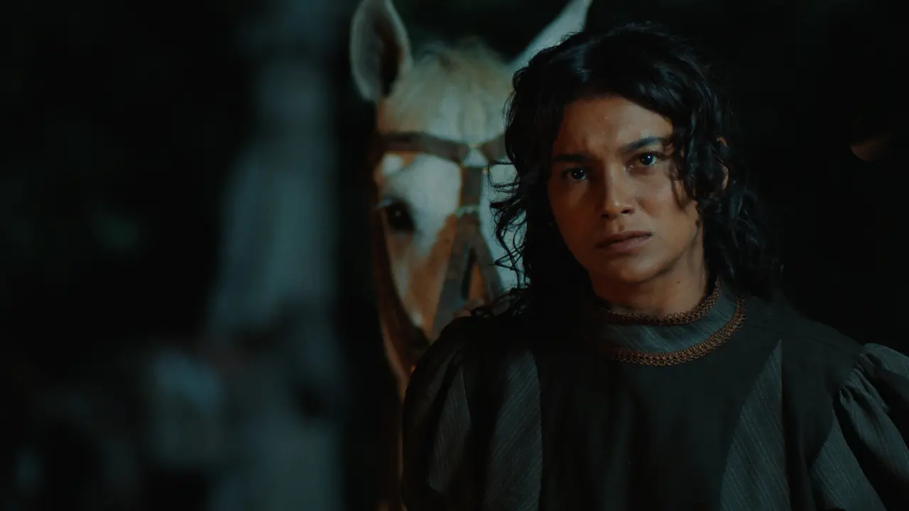 Foto escura e dramática da atriz Dira Paes, de cabelos longos e escuros, no filme 'Lua Cambará'. Ela está vestindo uma roupa de época escura com um detalhe de gola dourada, olhando para a frente com uma expressão séria. Ao fundo, fora de foco e ligeiramente desfocado, está a cabeça de um cavalo branco ou de cor clara. A iluminação é baixa, criando uma atmosfera noturna ou sombria.