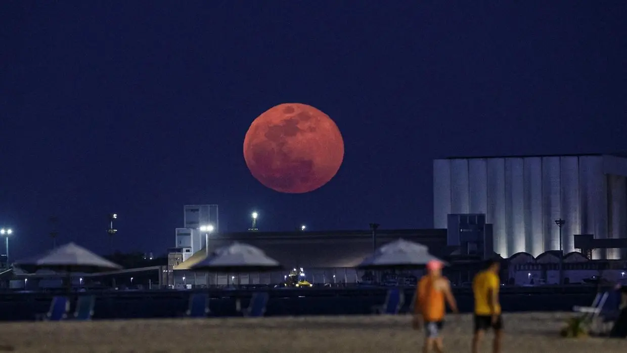 Imagem noturna de uma grande lua cheia avermelhada sobre a orla de Fortaleza, capital do Ceará, com guarda-sóis, cadeiras e pessoas e edifícios ao fundo.
