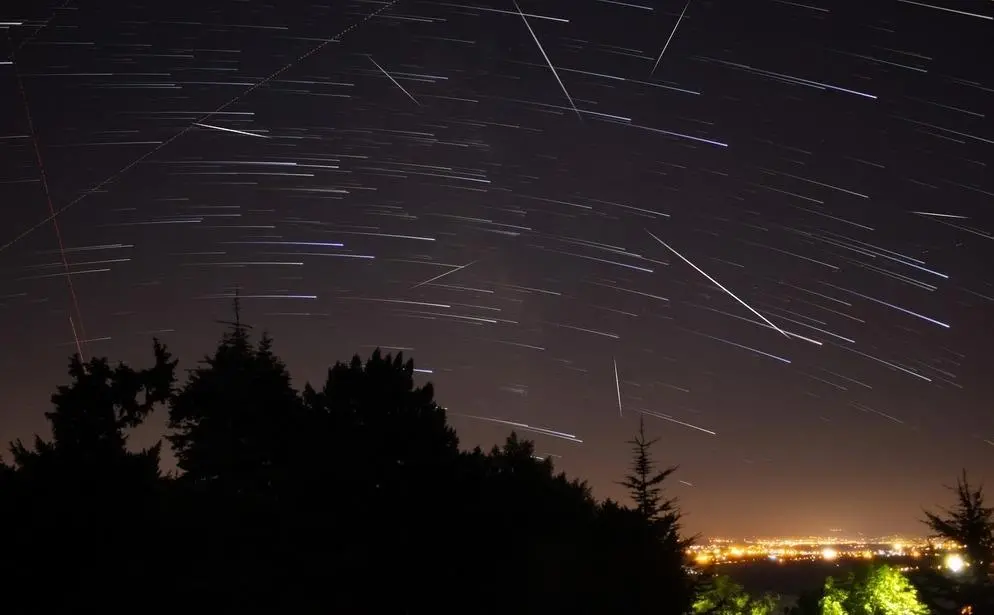 Imagem mostra céu noturno com rastros luminosos de estrelas e vários meteoros cadentes, sobre a silhueta de árvores e luzes distantes de uma cidade.
