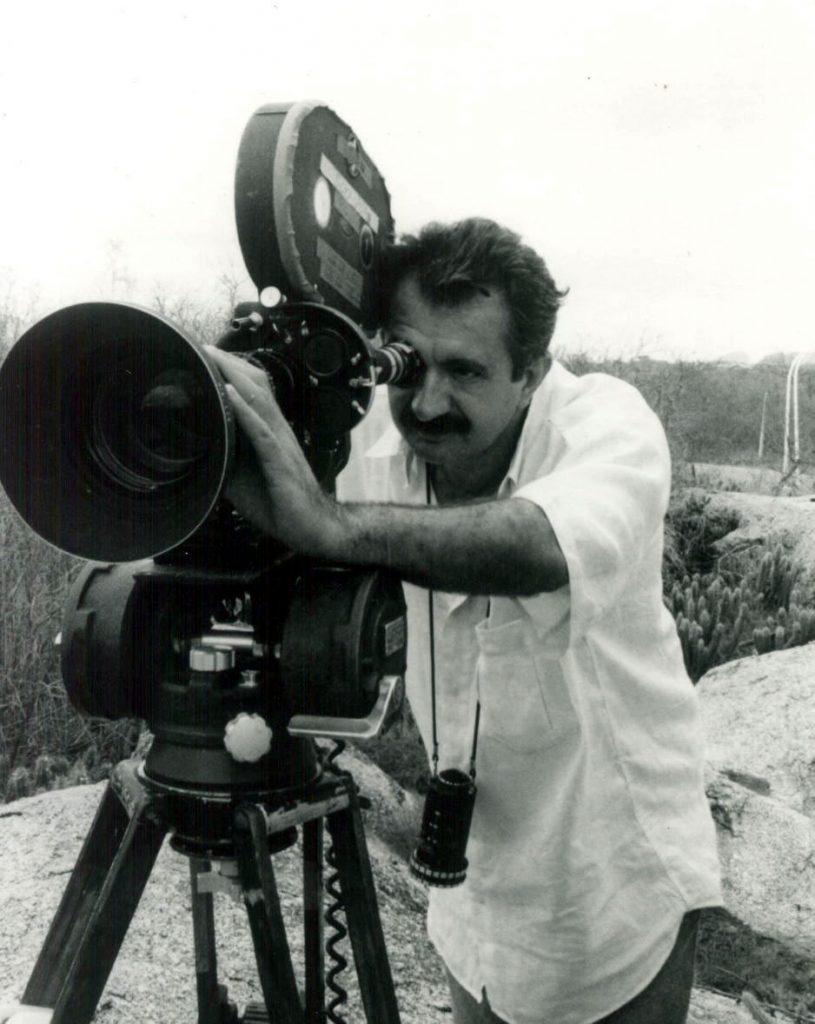 Foto em preto e branco do cineasta Rosemberg Cariry, que está de blusa branca e usa uma câmera de cinema.