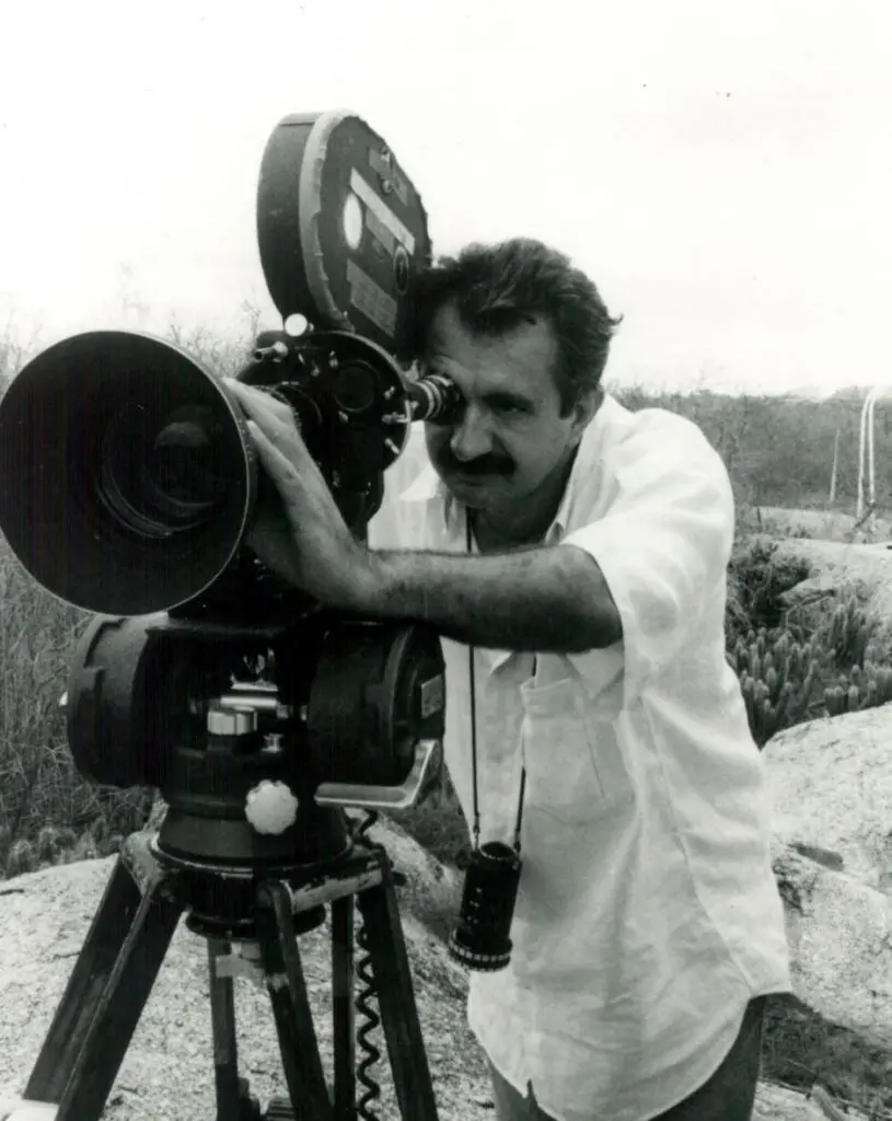 Foto em preto e branco do cineasta Rosemberg Cariry, que está de blusa branca e usa uma câmera de cinema.