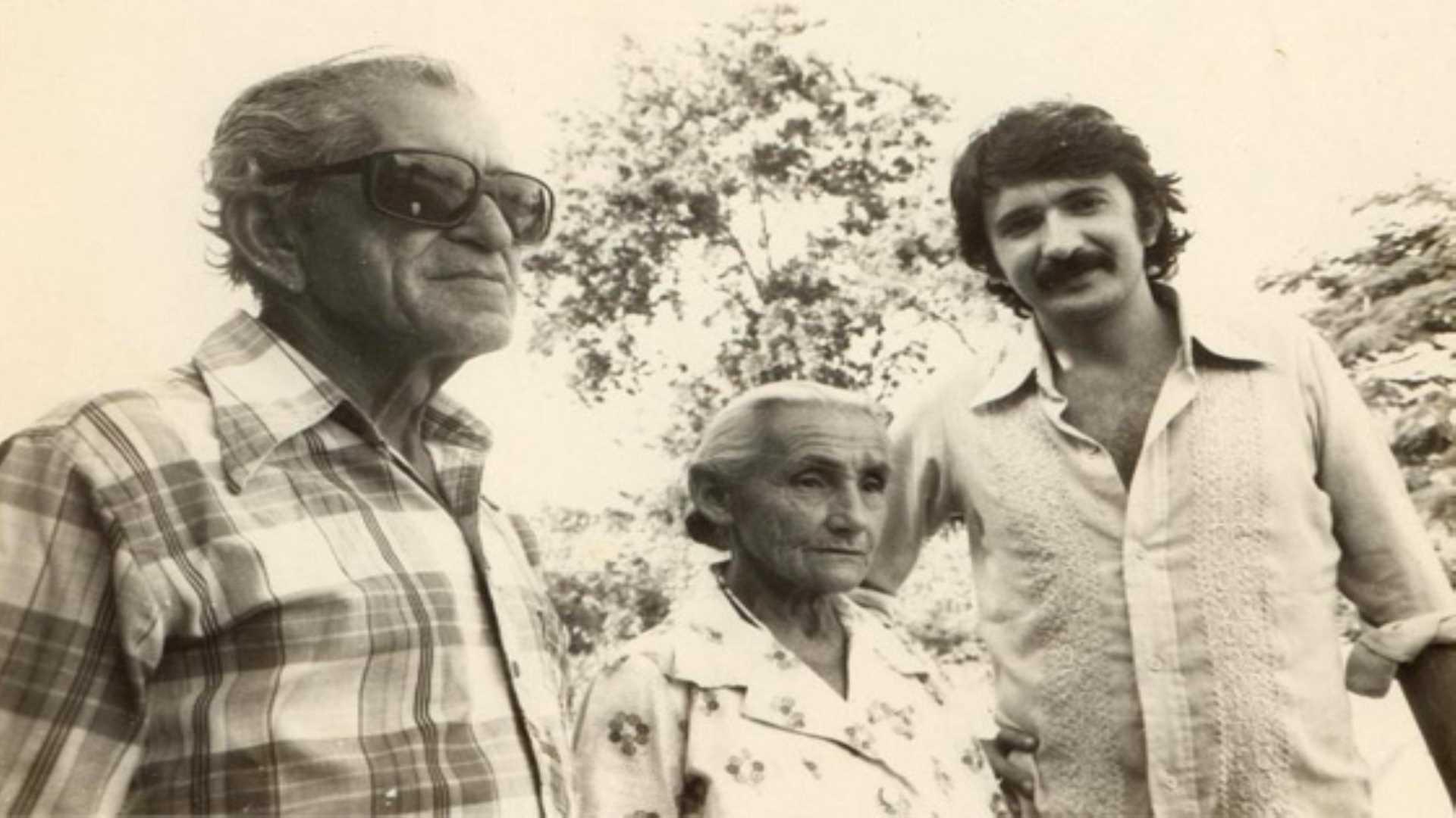 Uma fotografia em preto e branco (ou sépia clara) mostra três pessoas em um ambiente externo, com folhagem e árvores desfocadas ao fundo. À esquerda, um homem idoso, Patativa do Assaré, usa óculos de sol escuros e uma camisa xadrez de manga curta. Ao centro, uma mulher idosa, Dona Belinha (esposa de Patativa), está entre os dois homens. Ela tem cabelos claros presos, usa uma blusa estampada e está olhando diretamente para frente. À direita, um homem mais jovem, Rosemberg Cariry, com bigode escuro e cabelo na altura da orelha, usa uma camisa clara abotoada.