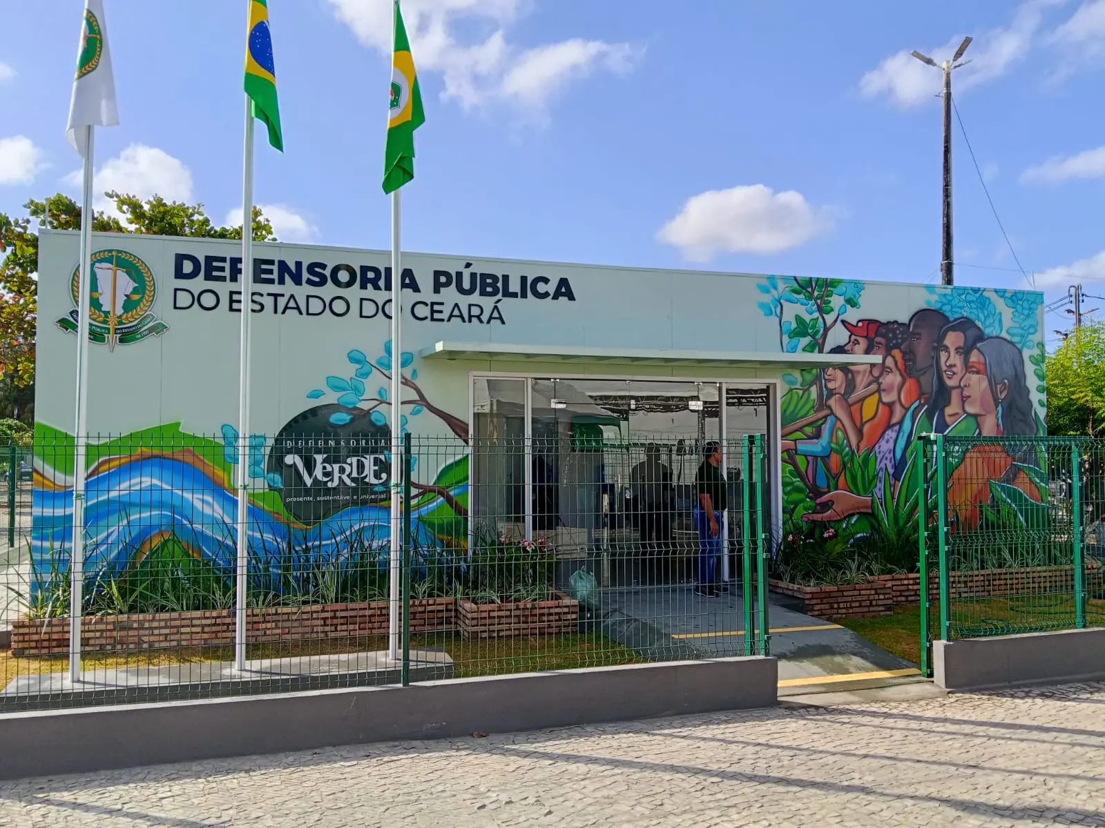 Fachada de uma unidade da Defensoria Pública do Estado do Ceará, com o letreiro institucional e o logotipo na parede. O prédio tem pintura colorida com elementos naturais e um mural artístico representando pessoas de diferentes etnias e gêneros em meio a vegetação verde. Na frente, há três mastros com bandeiras hasteadas — do Brasil, do Ceará e da Defensoria Pública. O local é cercado por grade verde e possui calçada de paralelepípedos. O céu está azul com algumas nuvens.