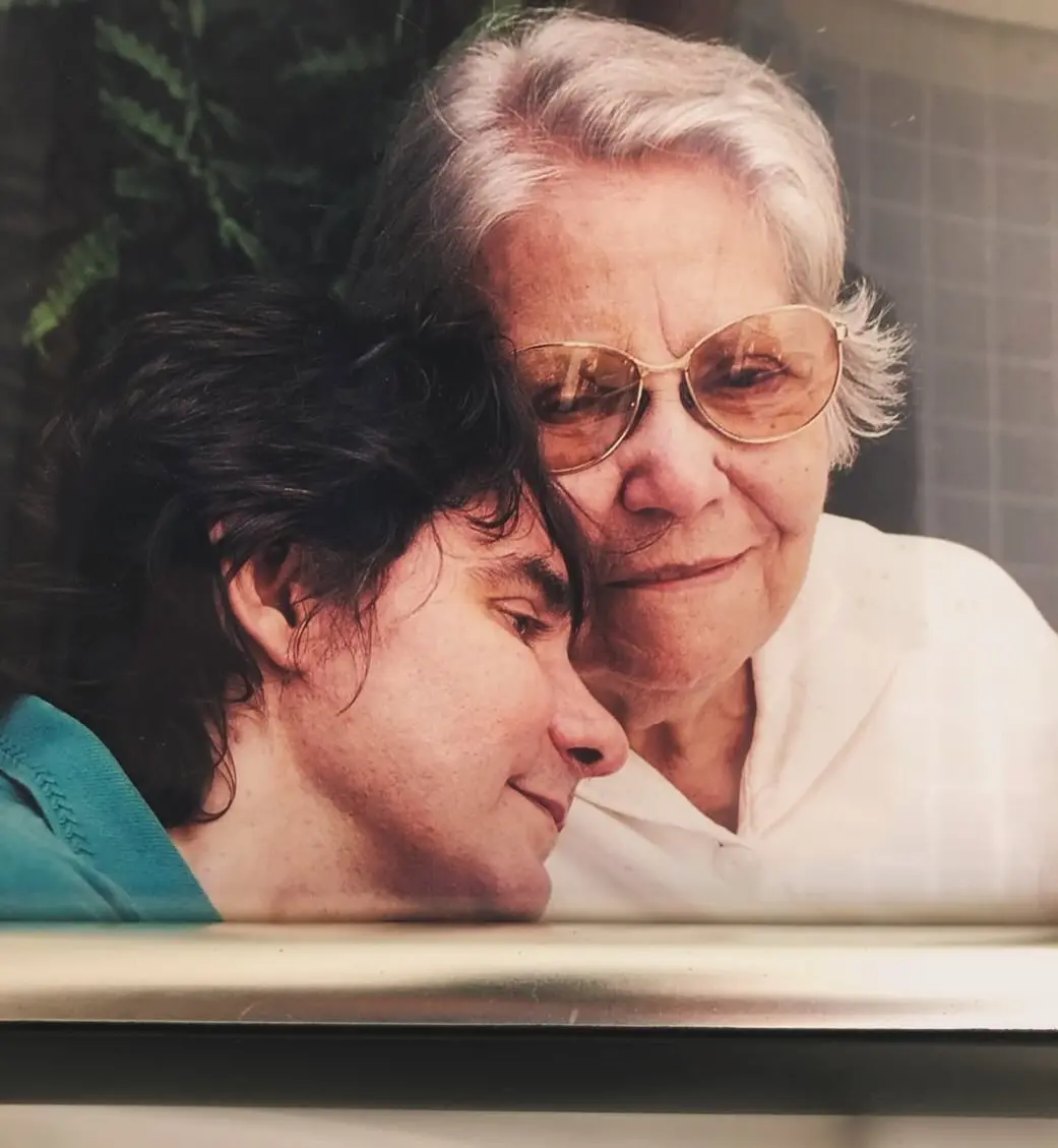 Na imagem, uma foto em close-up, possivelmente emoldurada, de duas pessoas juntas em um momento de carinho. No primeiro plano, à esquerda, está Lô Borges, com cabelo escuro e ombro em uma blusa azul-turquesa, encostando a cabeça gentilmente na outra pessoa. À direita e ligeiramente atrás, está uma senhora mais velha, Dona Maricota (mãe dos irmãos Borges), com cabelo branco e óculos de armação dourada com lentes claras. Ela veste uma blusa branca e olha com um sorriso sutil e afetuoso para a câmera ou para a frente. A imagem transmite intimidade e ternura.