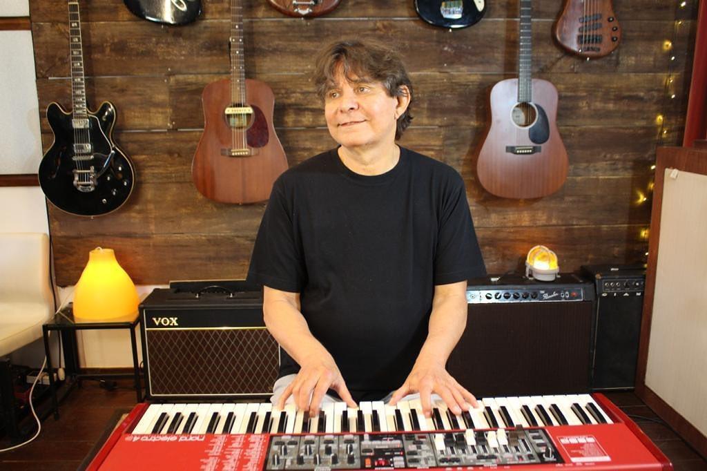Na imagem, Lô Borges, o músico, de meia-idade, com cabelo castanho-escuro, em um estúdio musical. Ele veste uma camiseta preta lisa e está sentado ou em pé atrás de um teclado sintetizador vermelho (Nord Electro), com as mãos sobre as teclas. Ele sorri sutilmente e olha para a esquerda (para fora da câmera). O fundo é uma parede de madeira rústica e escura, onde várias guitarras e violões de diferentes estilos e cores estão pendurados. Equipamentos musicais, como amplificadores (um da marca VOX) e luminárias (incluindo uma laranja à esquerda), preenchem o espaço.