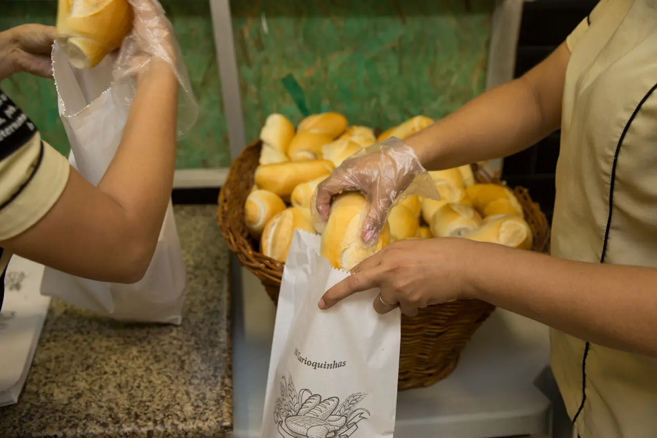 Pão carioquinha sendo colocado em saco de pão por mão de uma pessoa negra. Ao lado, outra mão de outra pessoa negra também colocando pão caroquinha em um saco.