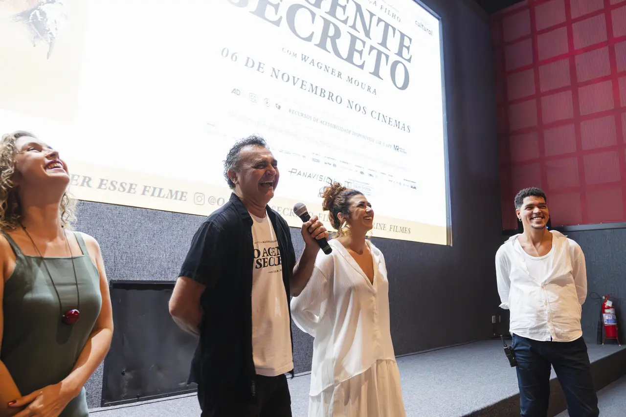 Quatro pessoas sorrindo no palco de um cinema durante a pré-estreia do filme 'O Agente Secreto'. No centro, Robério Diógenes, um homem com camisa preta e um microfone, ri, ao lado de uma mulher de roupa branca, Geane Albuquerque também sorrindo. À esquerda, Cely Farias, uma mulher de vestido verde, também rindo. À direita, Fabio Rodrigues Filho, um homem de camisa branca de botões desabotoada no pescoço e calça escura. A tela ao fundo exibe o título do filme e a data '06 de novembro nos cinemas'.