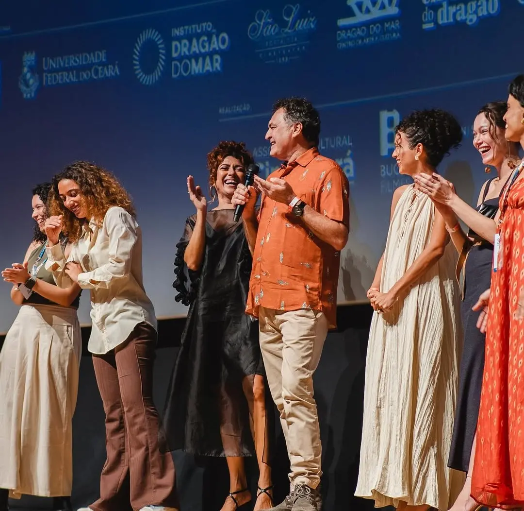 Um grupo de sete pessoas, incluindo os atores cearenses Geane Albuquerque (de vestido preto, ao centro) e Robério Diógenes (de blusa laranja estampada, segurando um microfone), no palco de um evento de cinema (35º Cine Ceará). O restante das pessoas está sorrindo e aplaudindo, trajando roupas casuais e elegantes. A tela ao fundo exibe logotipos de patrocinadores e instituições culturais cearenses.