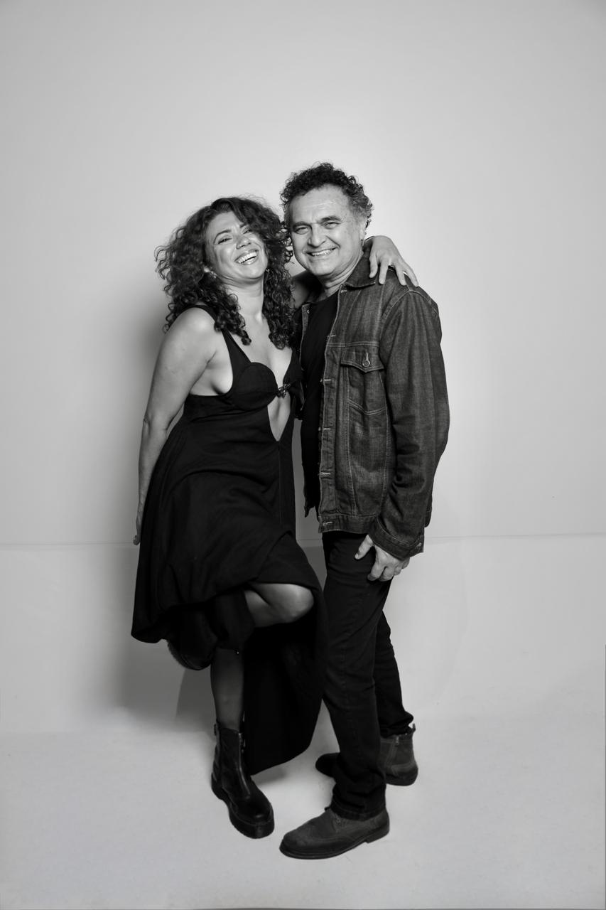 Retrato em preto e branco de Geane Albuquerque e Robério Diógenes. A mulher, à esquerda, sorri de forma vibrante, vestindo um vestido preto e botas. O homem, à direita, sorri levemente e veste jaqueta jeans e calças escuras, com o braço em volta do ombro dela. Ambos posam em frente a um fundo liso e claro.