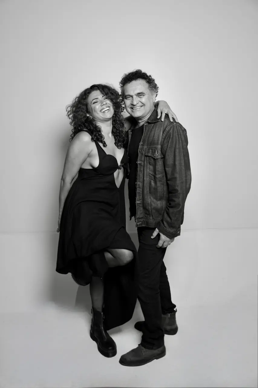 Retrato em preto e branco de Geane Albuquerque e Robério Diógenes. A mulher, à esquerda, sorri de forma vibrante, vestindo um vestido preto e botas. O homem, à direita, sorri levemente e veste jaqueta jeans e calças escuras, com o braço em volta do ombro dela. Ambos posam em frente a um fundo liso e claro.