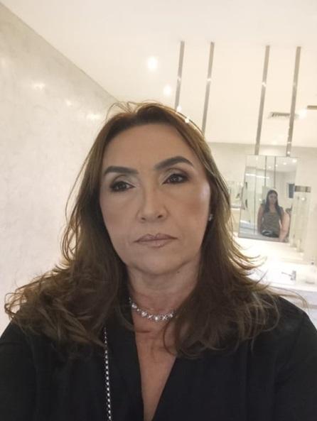 Na imagem, selfie de uma mulher de meia-idade, com cabelos castanhos ondulados e maquiagem sutil, olhando diretamente para a câmera. Ela usa uma blusa preta e um colar de pedras brancas em formato de 'X'. A foto foi tirada em um banheiro moderno, com paredes e bancada de mármore claro. No espelho atrás dela, pode-se ver o reflexo de outra mulher em uma blusa estampada.