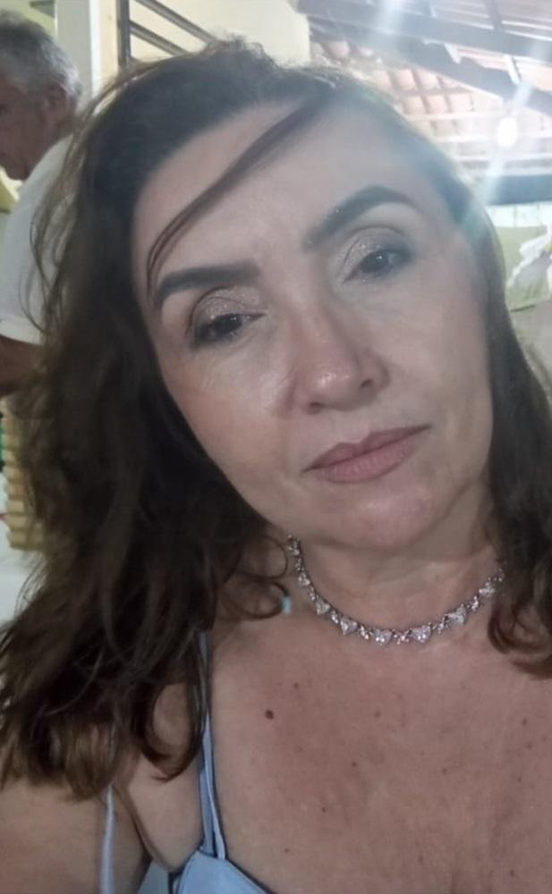 Na imagem, selfie em close-up de Idalina Mesquita, conhecida por sua semelhança com a escritora Clarice Lispector. Ela é uma mulher de meia-idade, com cabelos castanhos ondulados caindo sobre a testa. Ela usa maquiagem cintilante nos olhos, um vestido ou blusa de alça fina na cor azul clara e um colar brilhante com pingentes em formato de coração. No canto superior esquerdo, parte do ombro e cabelo de um homem idoso em uma camisa branca pode ser vista no fundo, que é levemente desfocado e iluminado.