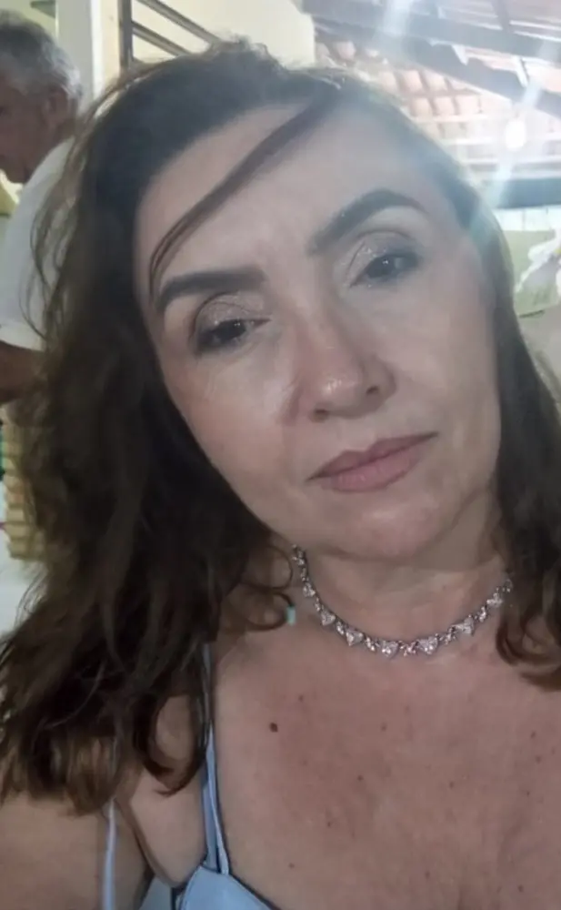 Na imagem, selfie em close-up de Idalina Mesquita, conhecida por sua semelhança com a escritora Clarice Lispector. Ela é uma mulher de meia-idade, com cabelos castanhos ondulados caindo sobre a testa. Ela usa maquiagem cintilante nos olhos, um vestido ou blusa de alça fina na cor azul clara e um colar brilhante com pingentes em formato de coração. No canto superior esquerdo, parte do ombro e cabelo de um homem idoso em uma camisa branca pode ser vista no fundo, que é levemente desfocado e iluminado.