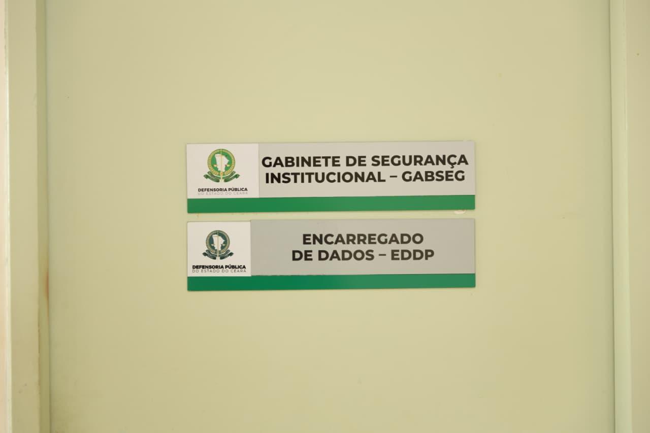 Placas afixadas em uma porta de cor clara, com logotipos da Defensoria Pública do Estado do Ceará. A placa superior tem o texto “Gabinete de Segurança Institucional – GABSEG” e a inferior diz “Encarregado de Dados – EDDP”. Ambas possuem uma faixa verde na parte inferior.