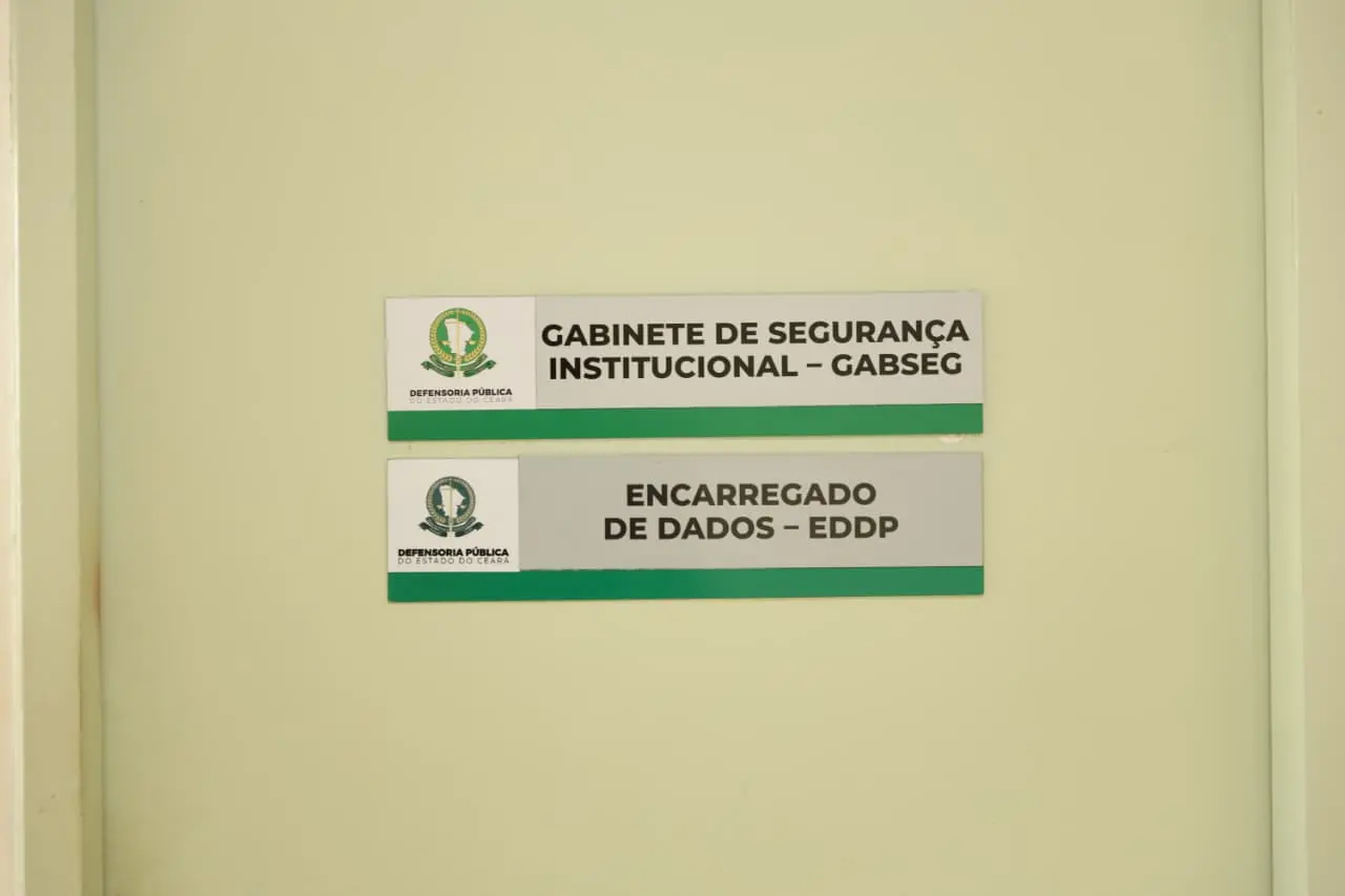 Placas afixadas em uma porta de cor clara, com logotipos da Defensoria Pública do Estado do Ceará. A placa superior tem o texto “Gabinete de Segurança Institucional – GABSEG” e a inferior diz “Encarregado de Dados – EDDP”. Ambas possuem uma faixa verde na parte inferior.
