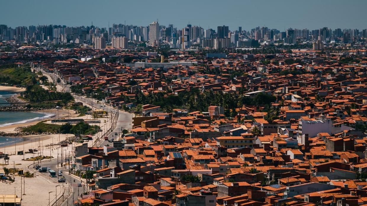 Vista aérea de bairro litorâneo em Fortaleza, com casas de telhados avermelhados, avenida costeira e mar à esquerda, e área urbana densa com prédios altos ao fundo sob céu claro.