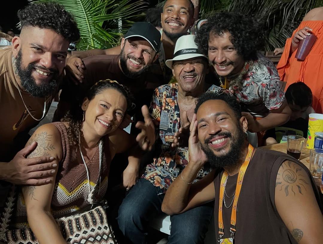 Foto de grupo alegre, noturna e de close-up. O sambista Jorge do Pagode, de chapéu e camisa estampada, está no centro, sorrindo e gesticulando, rodeado por seis membros do Grupo Mais Melanina e amigos. Todos estão sorrindo, celebrando um momento de convívio, possivelmente após ou durante uma apresentação na Barraca Foi Sol.
