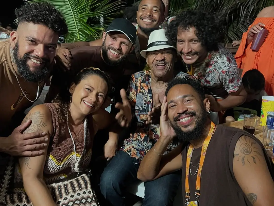 Foto de grupo alegre, noturna e de close-up. O sambista Jorge do Pagode, de chapéu e camisa estampada, está no centro, sorrindo e gesticulando, rodeado por seis membros do Grupo Mais Melanina e amigos. Todos estão sorrindo, celebrando um momento de convívio, possivelmente após ou durante uma apresentação na Barraca Foi Sol.