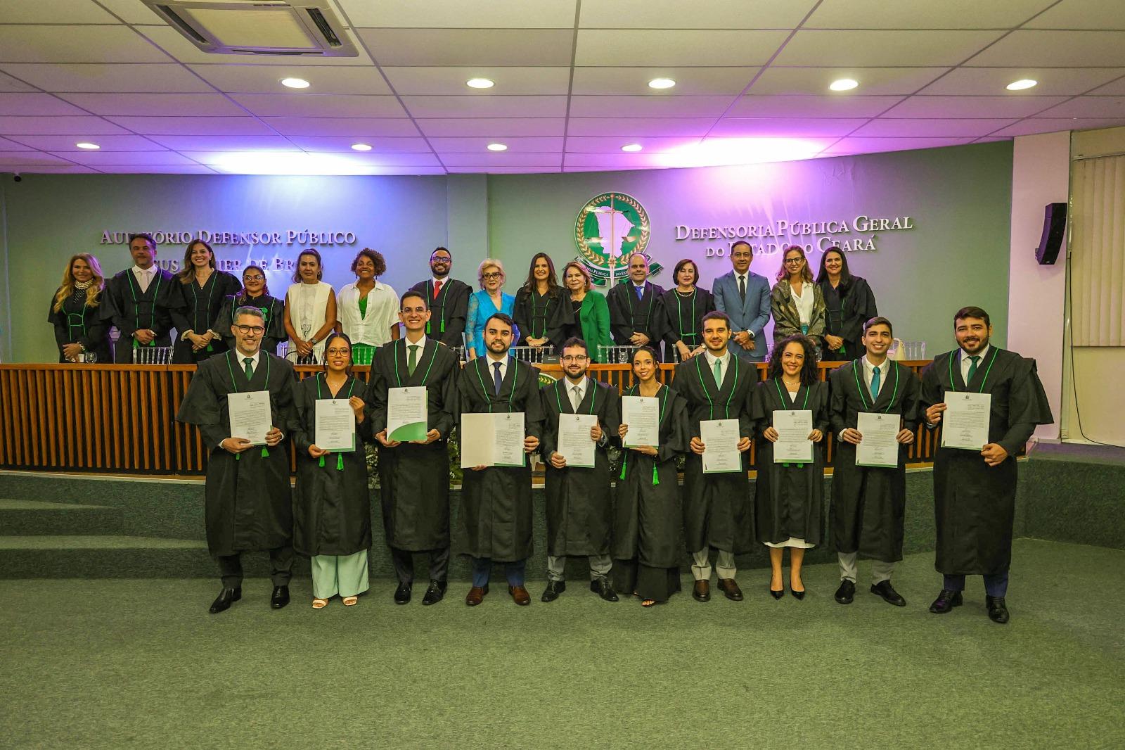 Grupo de novos defensores públicos do Estado do Ceará posa para foto oficial após cerimônia de posse. Na primeira fileira, dez pessoas vestidas com becas pretas e gravatas ou faixas verdes seguram seus certificados. Atrás deles, em um patamar elevado, há outras autoridades e servidores da Defensoria Pública, também em trajes formais. Ao fundo, na parede, lê-se “Defensoria Pública Geral do Estado do Ceará” e “Auditório Defensor Público José Maria de Barros Pinho”. O ambiente é um auditório iluminado, com tons de verde e luzes suaves.