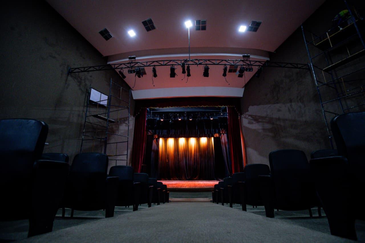 Na imagem, vista interna de um teatro, com perspectiva baixa do corredor central da plateia em direção ao palco. Fileiras de poltronas escuras estão alinhadas. O palco, ao fundo, é emoldurado por cortinas vermelhas e iluminado com luzes alaranjadas. Estruturas de andaimes são visíveis nas laterais, sugerindo um período de montagem ou manutenção.