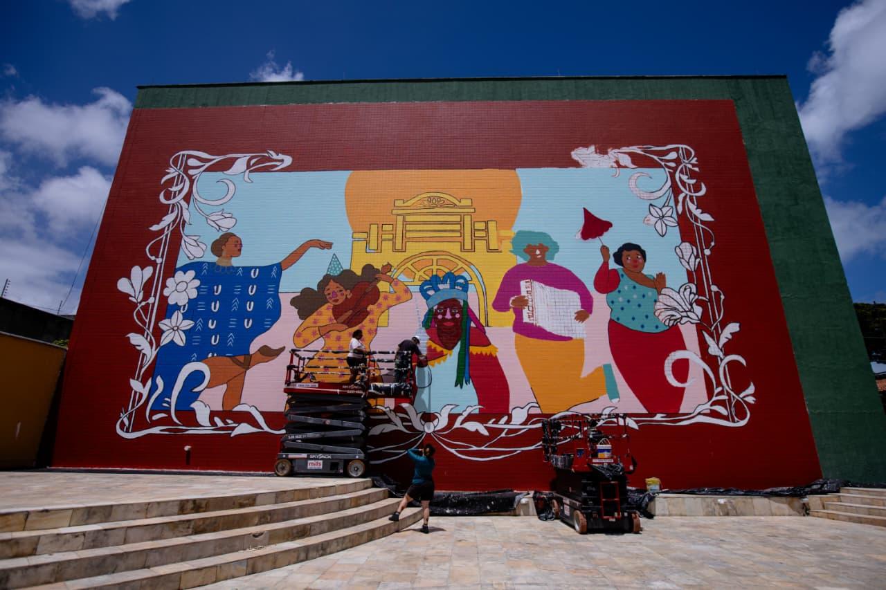 Na imagem, grande mural artístico colorido na parede externa de um prédio. O mural apresenta cinco figuras humanas estilizadas com roupas vibrantes, emolduradas por ornamentos brancos, com um sol e arquitetura ao fundo. Andaimes e equipamentos de trabalho estão visíveis na base, sugerindo que a pintura está em andamento. Degraus de pedra clara levam ao primeiro plano.