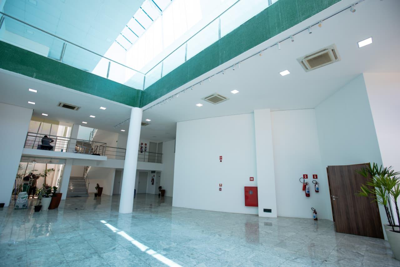 Na imagem, hall de entrada moderno e amplo com piso de mármore claro e paredes brancas. O espaço é iluminado por uma grande claraboia de vidro no teto e possui um mezanino com guarda-corpo de metal e vidro. Extintores de incêndio e plantas em vasos são visíveis nas laterais.