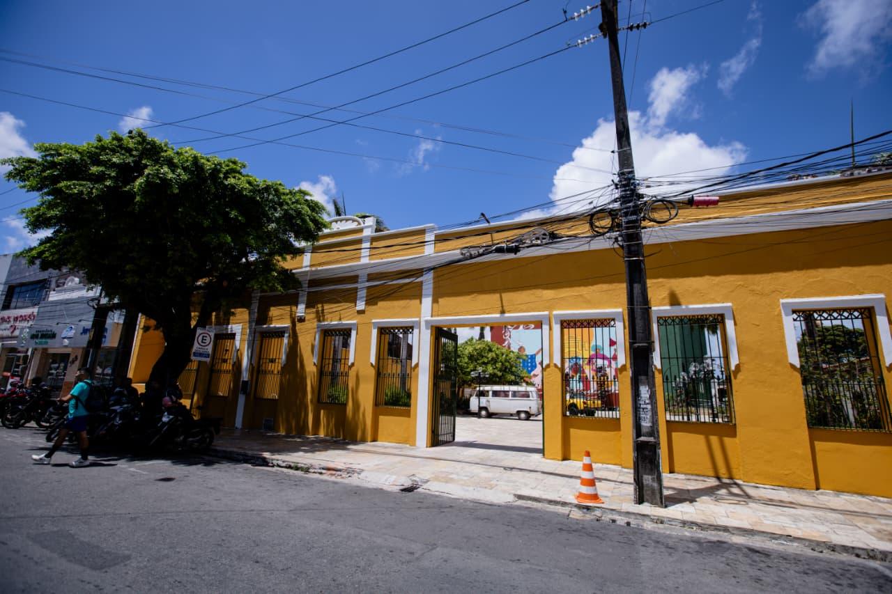 Na imagem, fachada de um prédio amarelo-ovo (Teatro Carlos Câmara) com janelas gradeadas, vista da rua. O portão central revela uma van branca e um mural colorido no pátio interno. Um poste de madeira com muitos cabos e um cone de sinalização está no primeiro plano à direita. Uma árvore e motocicletas estacionadas estão à esquerda.