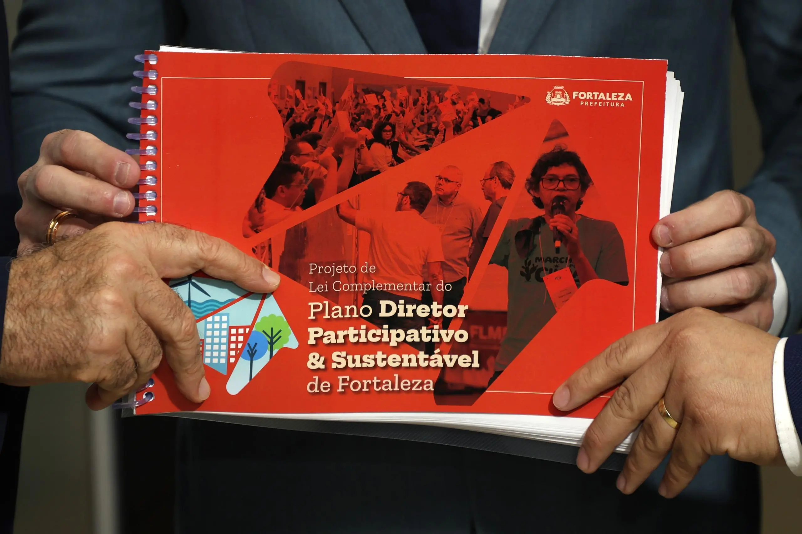 Livro encadernado, de cor laranja, com capa que anuncia se tratar do plano diretor de fortaleza, segurado por quatro mãos.