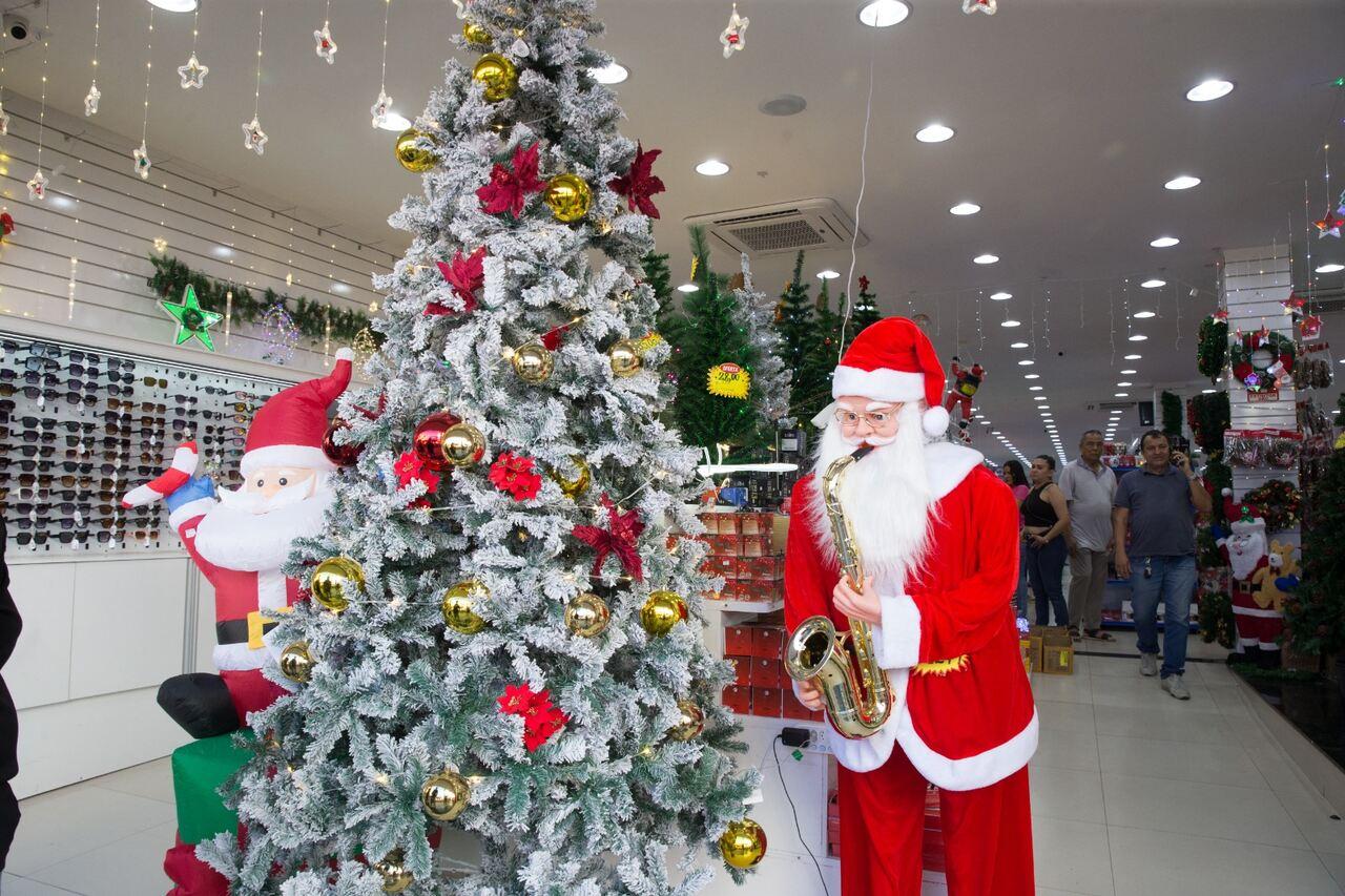 Árvore de galhos brancos e decoração dourada e vermelha em frente de loja de Natal. Ao lado esquerdo, um papai noel de tecido e, ao lado direito, um papai noel em tamanho real com um saxofone amarelo na boca.
