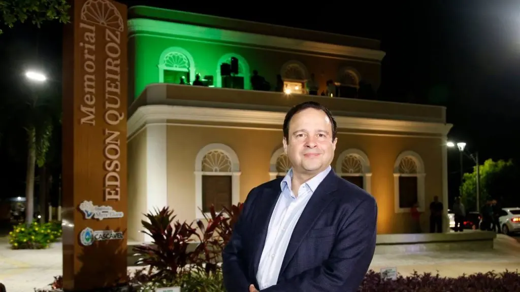 Retrato noturno de Igor Queiroz Barroso, um homem de terno azul-marinho, que aparece sorrindo e posicionado à frente do Memorial Edson Queiroz em Cascavel. O memorial tem a fachada iluminada em amarelo e o terraço superior em verde. Um totem vertical com o nome está visível à esquerda.