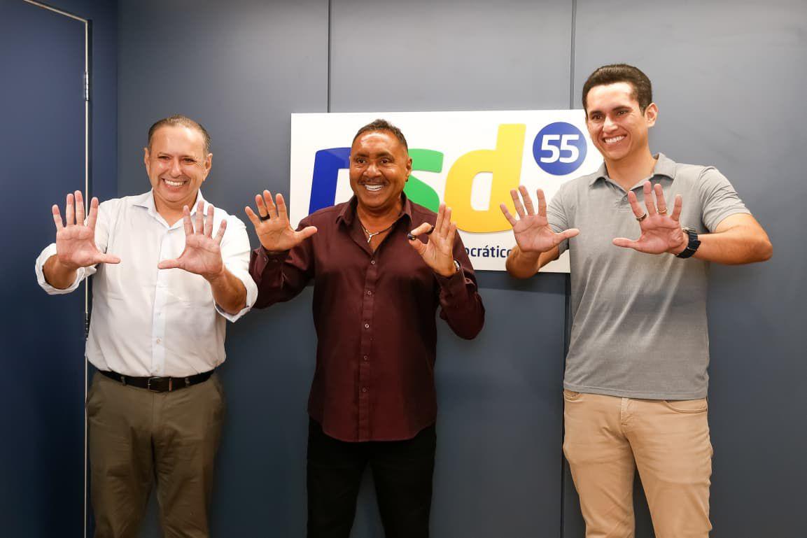Três homens posam sorrindo em frente a um painel com o logotipo do partido PSD e o número 55. Eles estão lado a lado, com as mãos levantadas mostrando os dedos abertos. O homem ao centro veste camisa vinho, o da esquerda usa camisa branca e calça cinza, e o da direita está com camisa polo cinza-clara e calça bege. O fundo é azul-acinzentado.