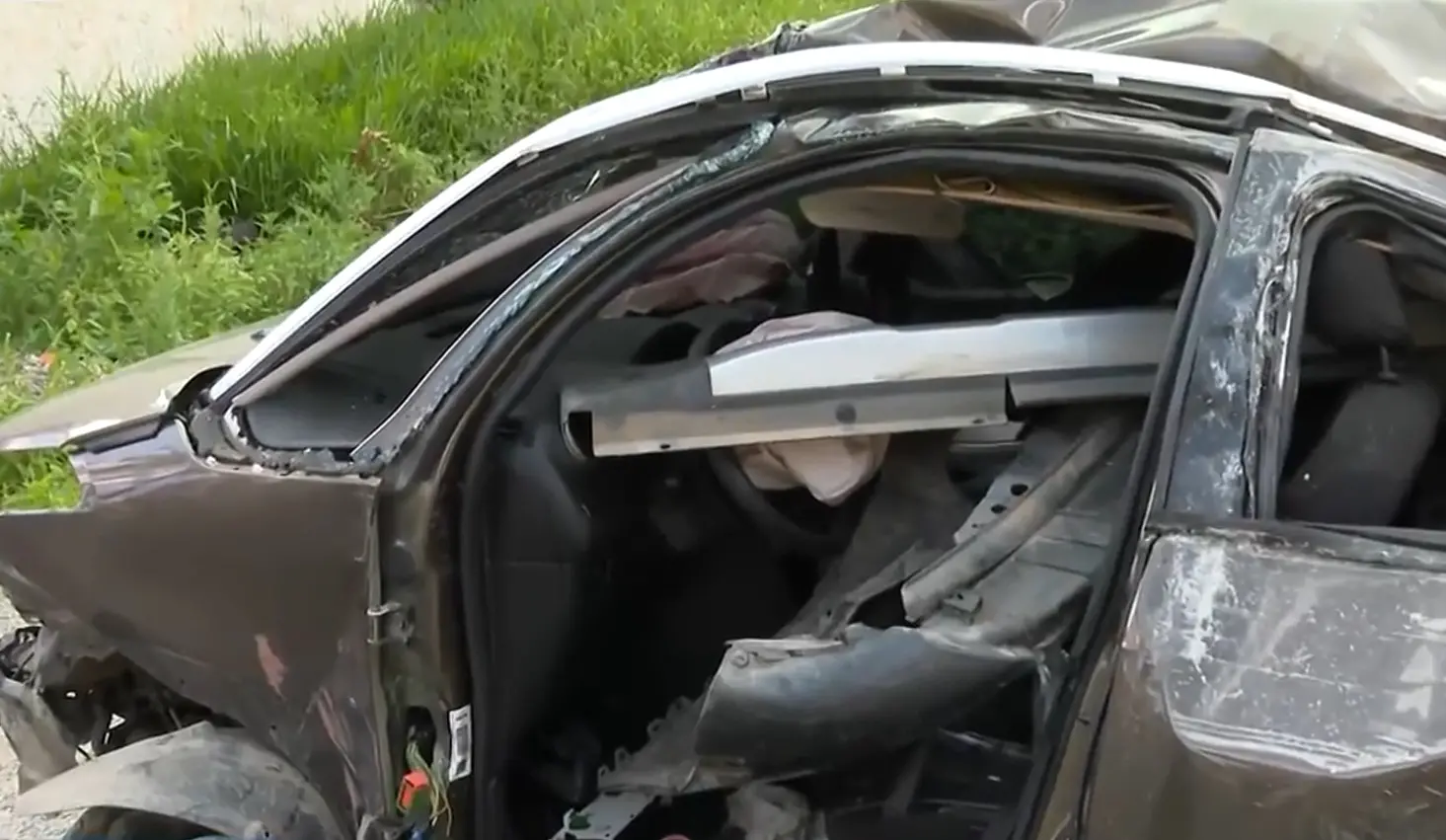 Imagem em close de um carro severamente danificado após um acidente, com o interior exposto e a estrutura do teto e das portas visivelmente destruída.