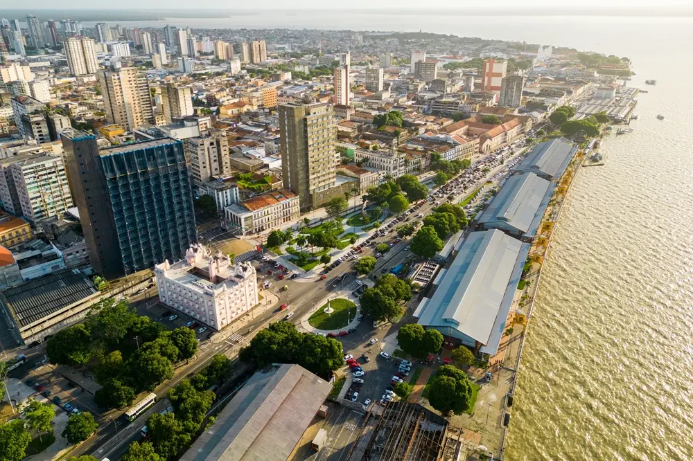 Na imagem, uma vista aérea de Belém, Pará, mostrando a Estação das Docas (estruturas longas e paralelas com telhados metálicos) ao longo da orla do rio. Em primeiro plano, há uma mistura de edifícios altos modernos e históricos, além de áreas arborizadas, praças e ruas movimentadas com carros. A água do rio à direita reflete o sol.