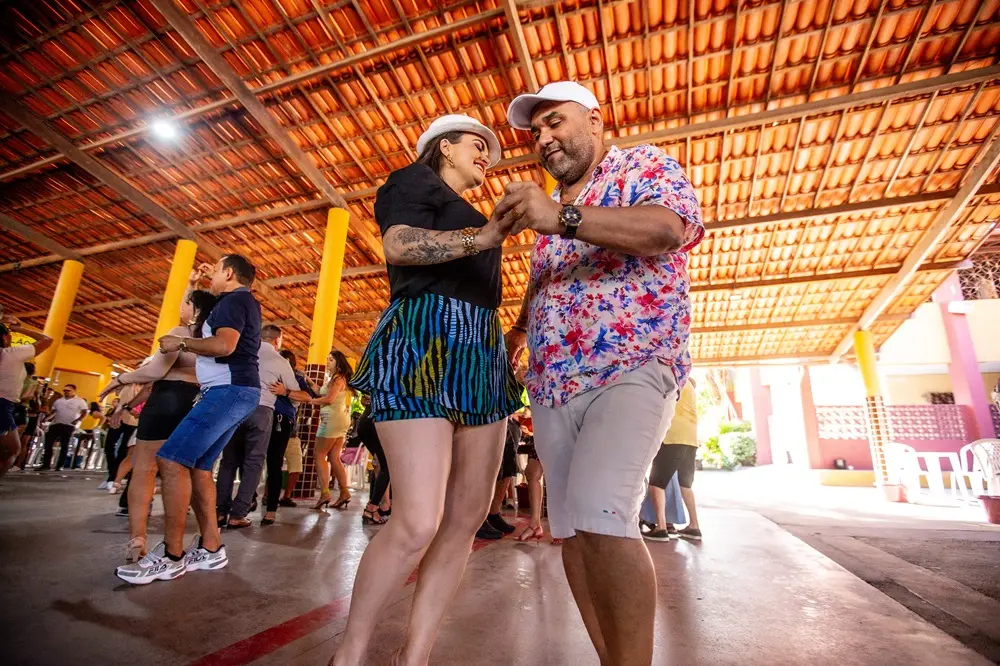 Na imagem, um casal de dançarinos em primeiro plano em um local de eventos aberto e coberto, com piso de concreto e teto de telhas cerâmicas. A mulher usa um chapéu fedora branco, uma blusa preta e um short colorido estampado. O homem veste um boné branco, uma camisa floral colorida e shorts cáqui. Eles estão de mãos dadas enquanto dançam. Várias outras pessoas estão dançando ao fundo.