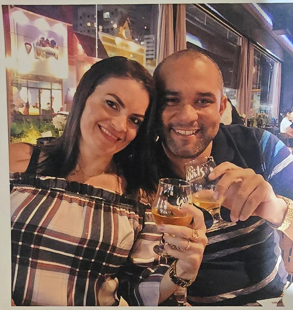 Na imagem, foto de close-up de um casal sorridente em um ambiente interno, provavelmente um restaurante ou bar, com iluminação suave. A mulher tem cabelos escuros e veste uma blusa xadrez de ombros nus em tons de azul marinho, branco e vermelho. O homem está ao lado dela, vestindo uma camisa polo escura com listras horizontais e um relógio de pulso dourado. Ambos seguram e brindam com pequenos copos de vidro contendo uma bebida âmbar. O fundo está desfocado com luzes noturnas e reflexos de vidro.