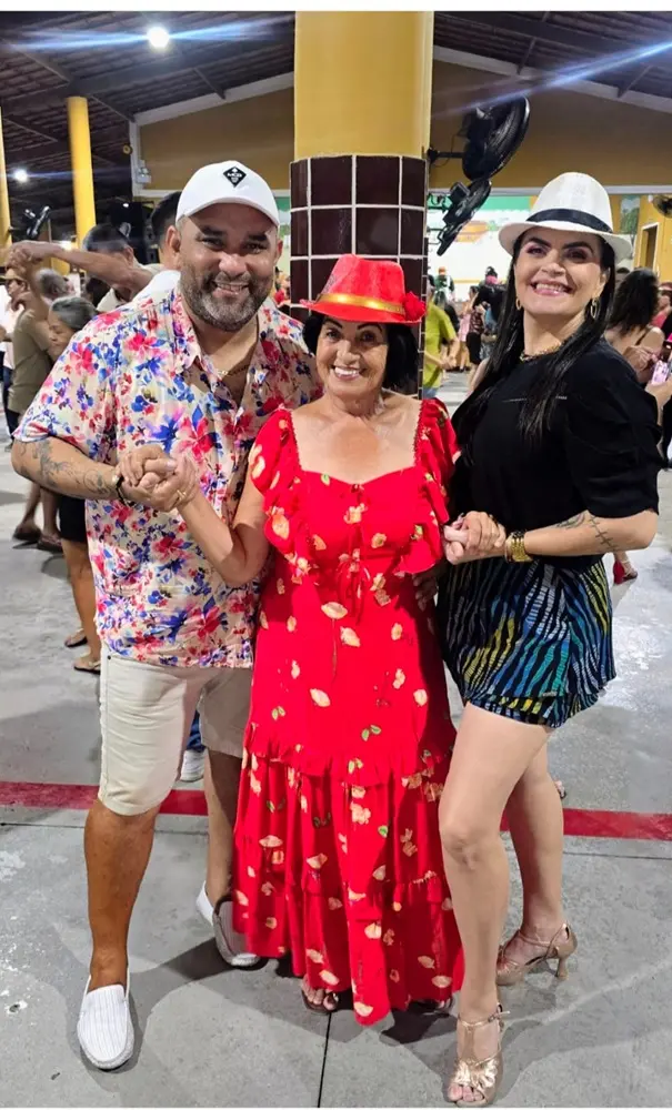 Na imagem, três pessoas sorrindo posam juntas em um ambiente de festa ou dança coberto. No centro, uma mulher mais velha sorri, vestindo um vibrante vestido vermelho com babados e um pequeno chapéu vermelho com uma fita. À esquerda, um homem com um boné branco e uma camisa floral colorida segura a mão dela. À direita, uma mulher com um chapéu fedora branco, blusa preta e shorts estampados coloridos segura a outra mão da mulher do centro. Eles estão em um piso de concreto com pessoas e pilares amarelos no fundo.