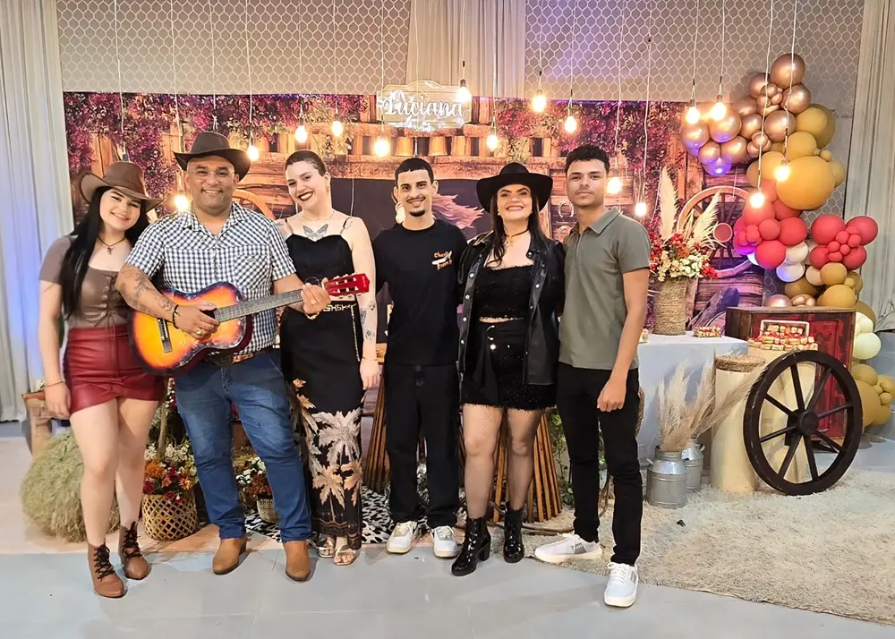 Na imagem, um grupo de seis pessoas posa para uma foto em um evento com tema rústico ou 'country'. Da esquerda para a direita: uma jovem de chapéu e saia de couro vinho; um homem de chapéu, camisa xadrez, jeans e botas, segurando um violão; uma mulher de vestido longo preto e gargantilha; um homem de camisa preta de manga curta; uma mulher de chapéu preto, jaqueta de couro preta e shorts; e um jovem de camisa polo verde oliva e calça escura. O cenário atrás deles é decorado com um letreiro de luzes, tecidos florais, um arranjo de balões em tons de cobre e um carrinho de madeira de roda grande.