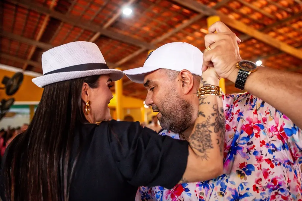 Na imagem, foto de close-up lateral de um casal dançando de rosto um para o outro em um local interno com teto de telhas cerâmicas. A mulher tem cabelo escuro liso e comprido, veste um chapéu fedora branco com uma faixa preta, brincos de argola dourados e uma blusa preta. O homem usa um boné branco, uma camisa floral colorida (tons de rosa, azul e branco), e exibe tatuagens e pulseiras douradas em seu braço. Ele também usa um relógio de pulso grande e dourado. Eles estão segurando as mãos acima da cabeça.