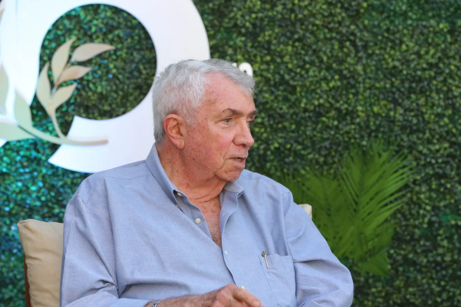 Homem branco de cabelo branco e blusa azul clara, sentado em uma cadeira de entrevista, com fundo de grama.