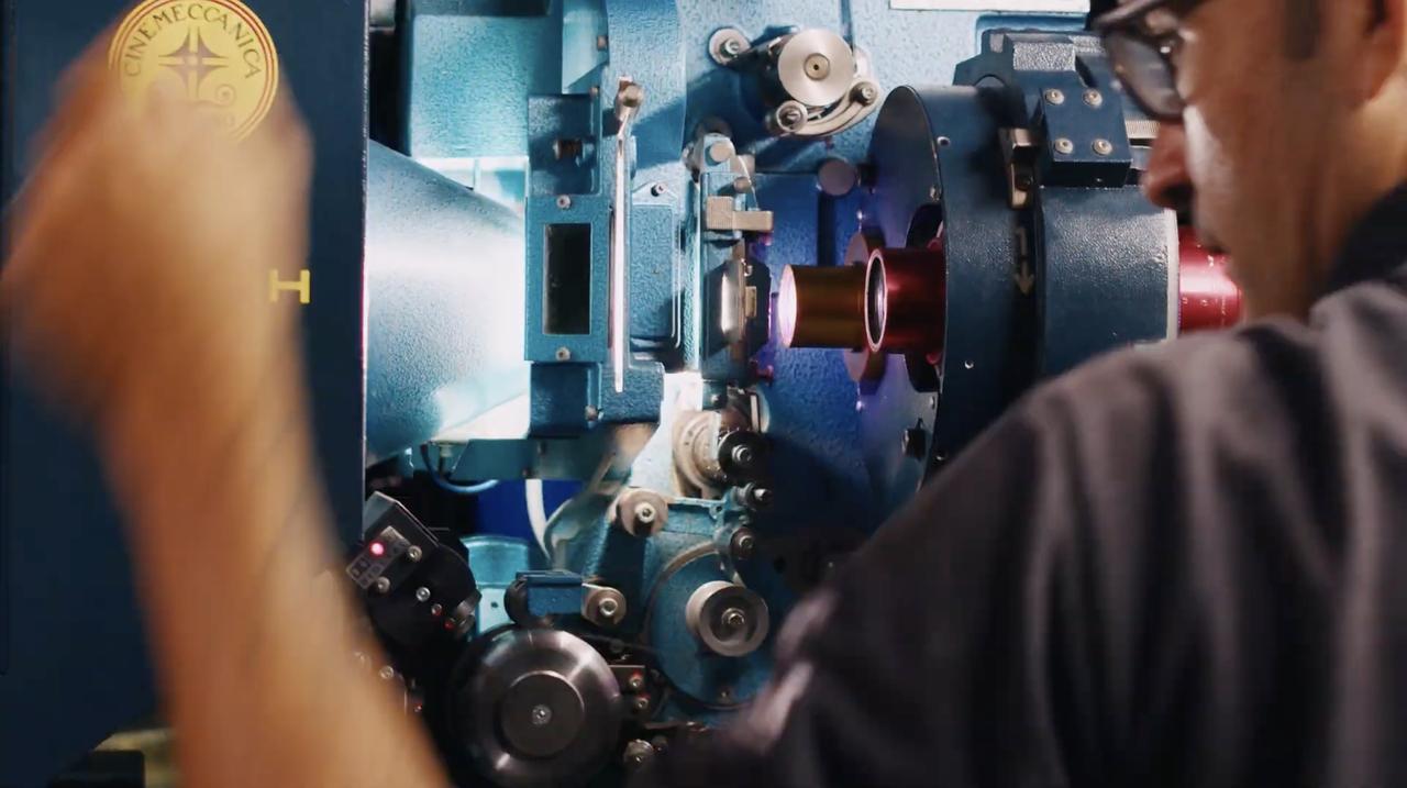 Um plano aproximado mostra um operador de cinema, visto de costas e em perfil, inspecionando o mecanismo interno de um projetor de cinema de cor azul-clara. O braço do operador, em movimento, está em primeiro plano à esquerda.