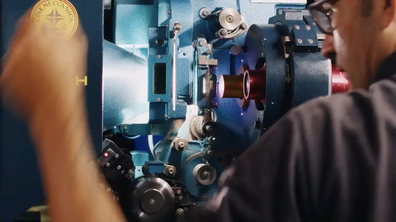 Um plano aproximado mostra um operador de cinema, visto de costas e em perfil, inspecionando o mecanismo interno de um projetor de cinema de cor azul-clara. O braço do operador, em movimento, está em primeiro plano à esquerda.