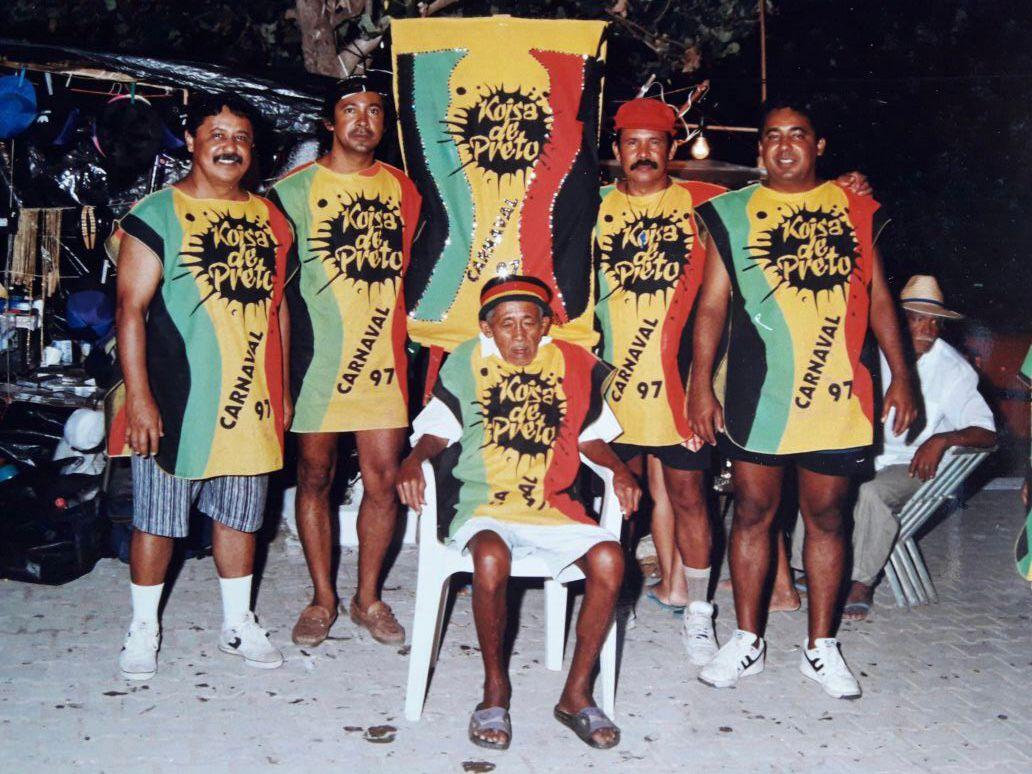 Uma fotografia colorida que captura um grupo de cinco homens posando para uma foto em um ambiente externo noturno, decorado com barracas à esquerda. Quatro homens estão em pé, lado a lado, enquanto um quinto está sentado em uma cadeira de plástico branca ao centro. Todos usam camisetas nas cores verde, amarelo, vermelho e preto com os dizeres 'Koisa de Preto - CARNAVAL 97'. Atrás do grupo, há um estandarte vertical com as mesmas cores e o nome do bloco. Um sexto homem, de chapéu de palha, está sentado em outra cadeira mais ao fundo, à direita.