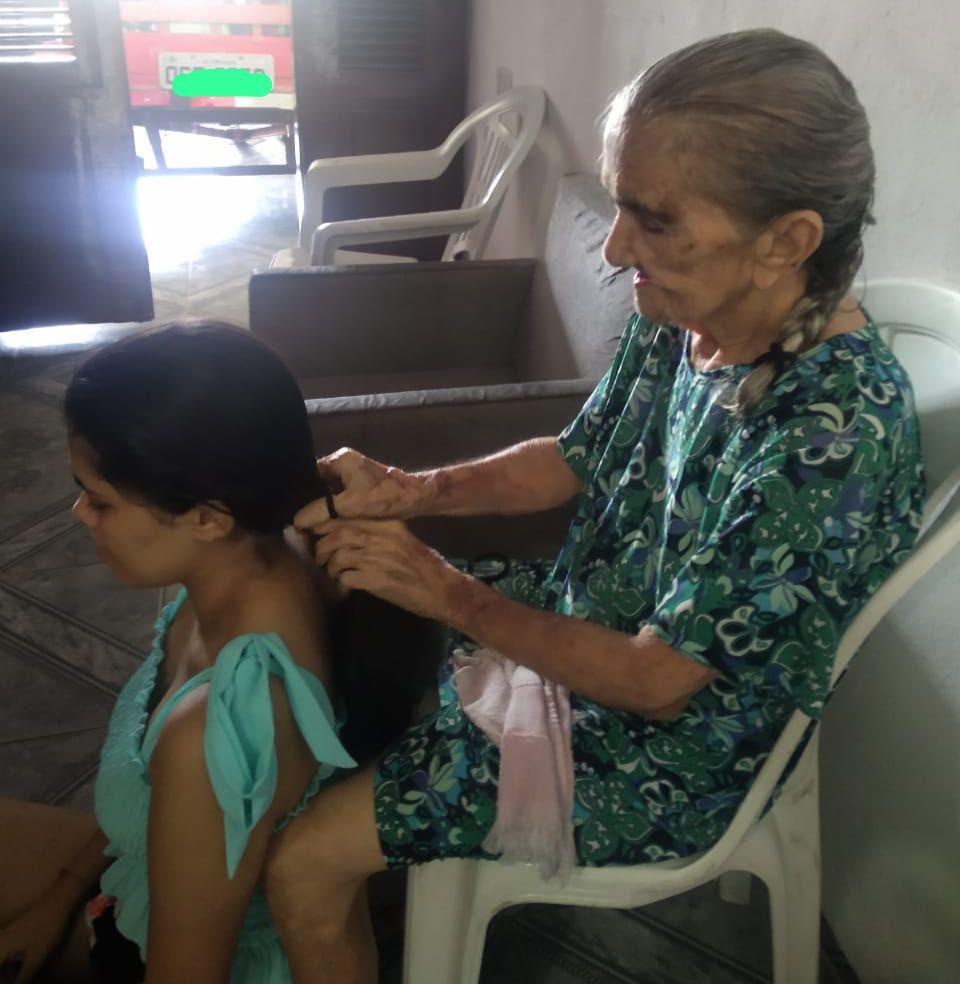 Idosa arrumando o cabelo de uma jovem em uma cadeira de plástico, em ambiente interno.